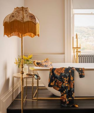 A bathroom with a white bathtub with a dark blue floral throw pillow and dressing gown in it, a tall yellow fringed lamp, white walls, and a window overlooking hills