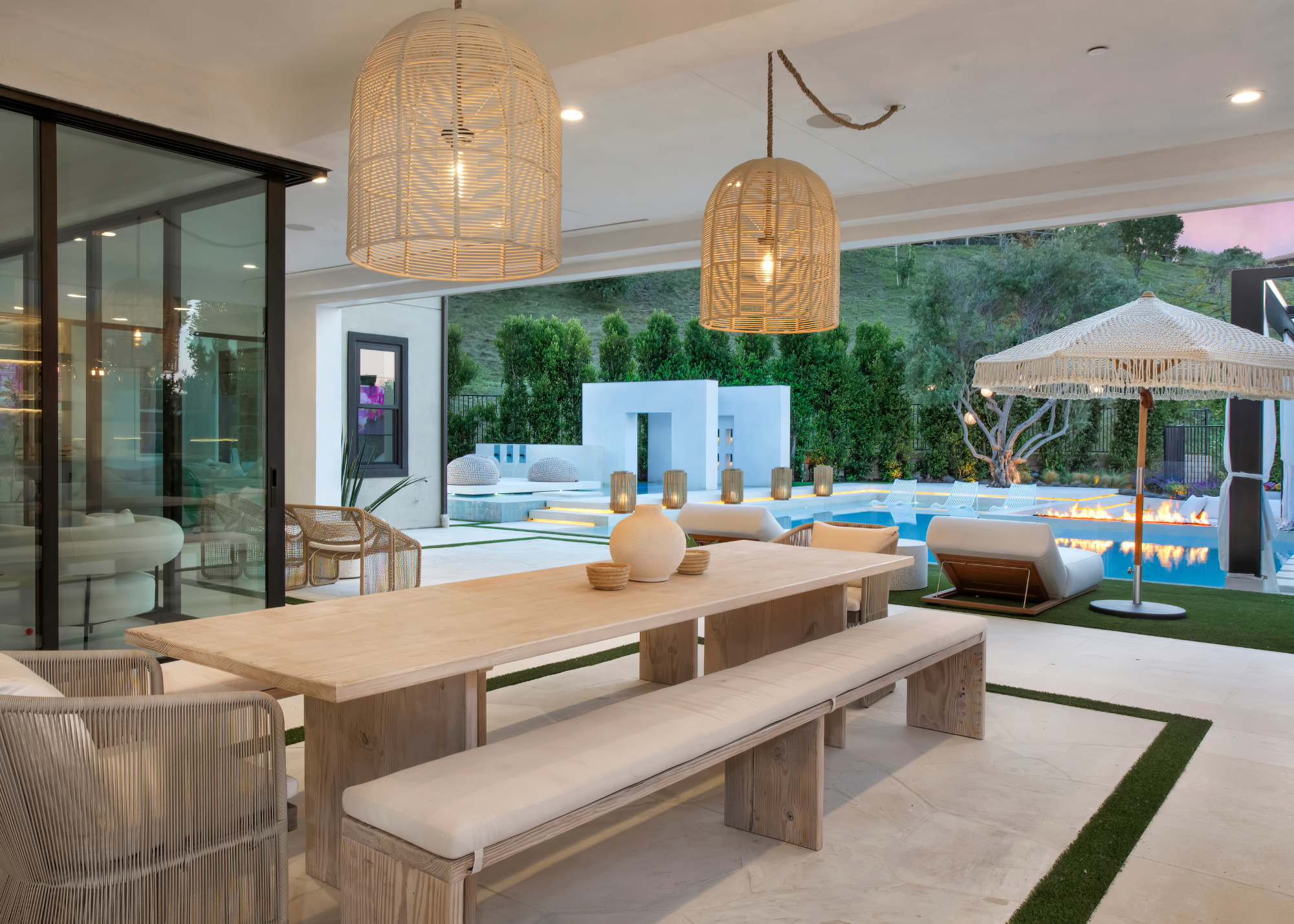 A modern Cali-cool indoor-outdoor area with a view overlooking a swimming pool; an outdoor dining bench with pendant lighting, lounge chairs, and a parasol with greenery in the distance to the side and beyond the pool
