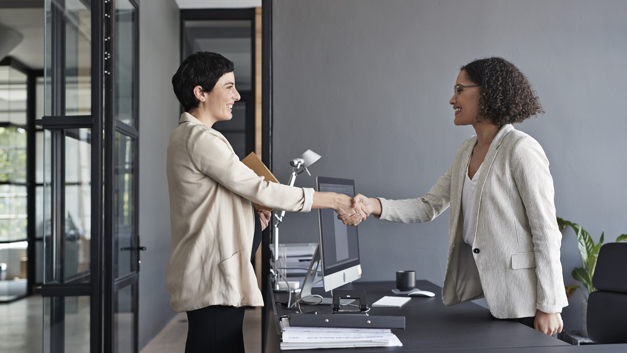 A woman shakes another woman's hand over her desk in a professional setting.