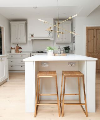 A kitchen with a white kitchen island with two wooden stools in frotn of it, a gold sputnik chandelier above it, and light gray cabinets and drawers behind it