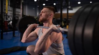 Man in a gym is squatting while lifting a barbell with heavy weights. He is visibly straining.