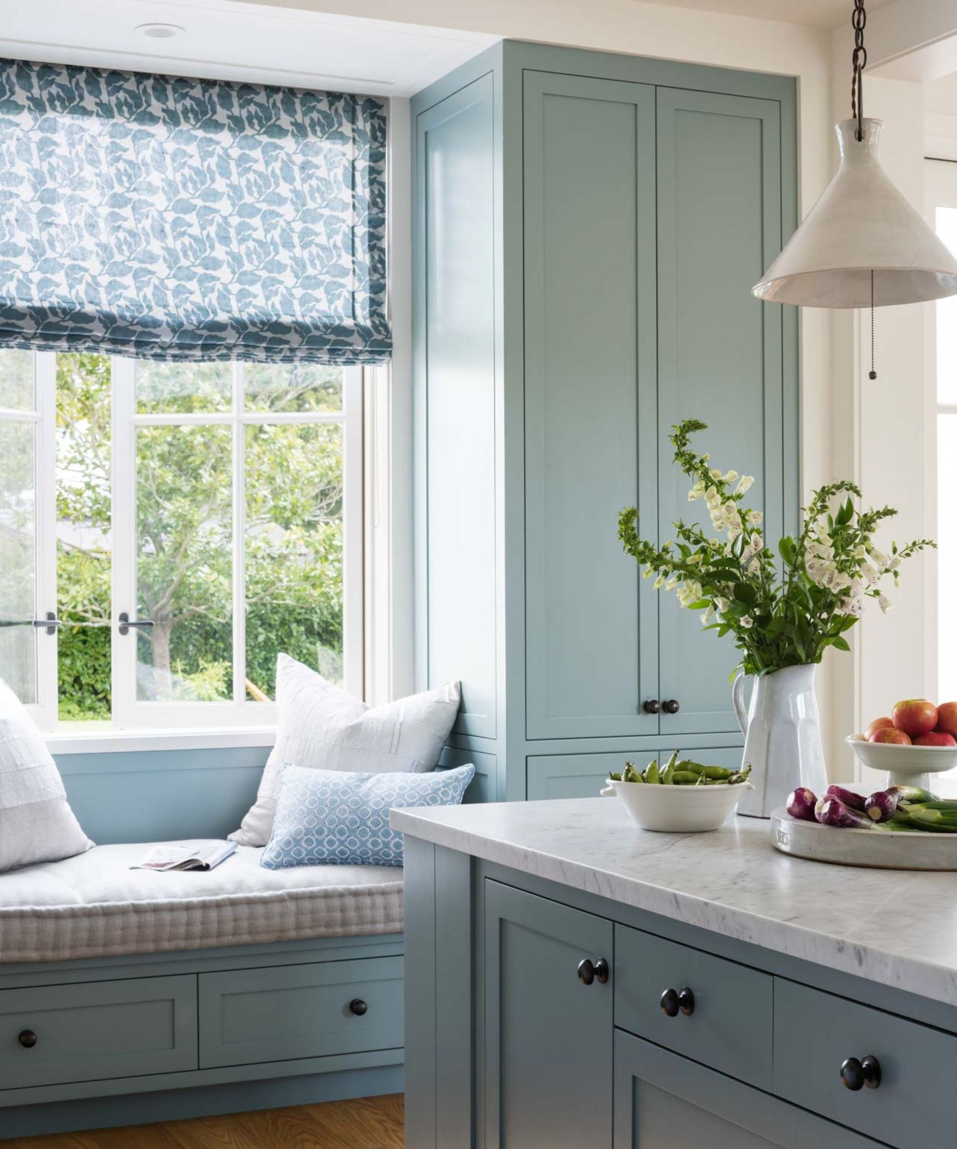 A midimalist kitchen with light blue cabinets, white marble countertops, a banquette window seat upholstered in gingham fabric, and a window dressed with a botanical blind