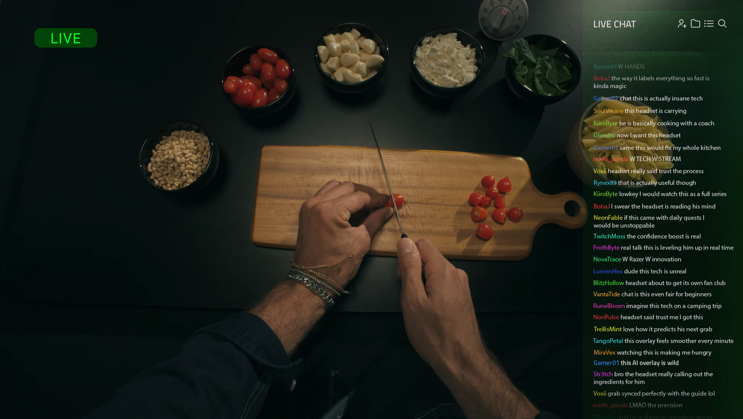 A top-down view of a person slicing cherry tomatoes on a wooden cutting board, surrounded by bowls of pine nuts, garlic cloves, crumbled cheese, and basil leaves.
