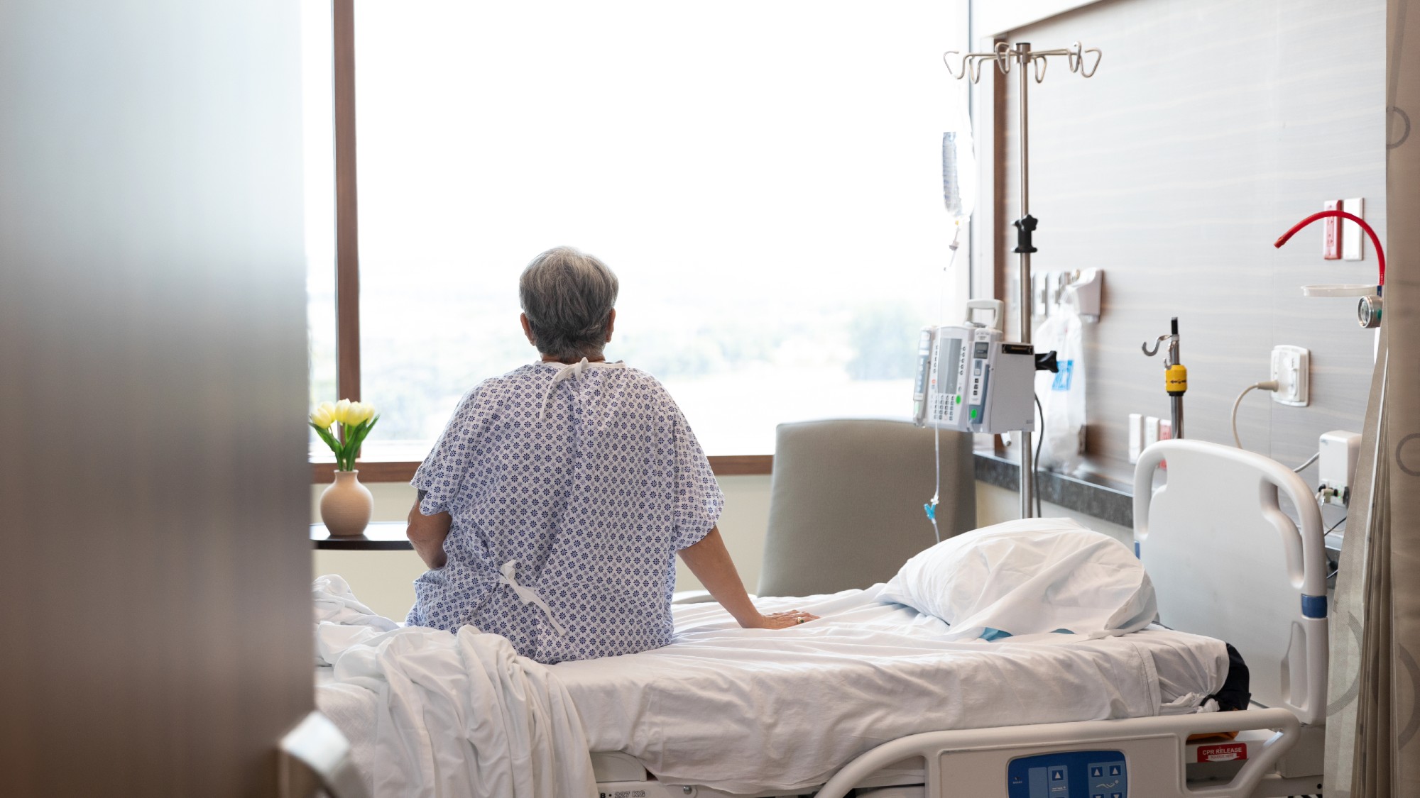 An elderly woman sits on a hospital bed with an IV drip