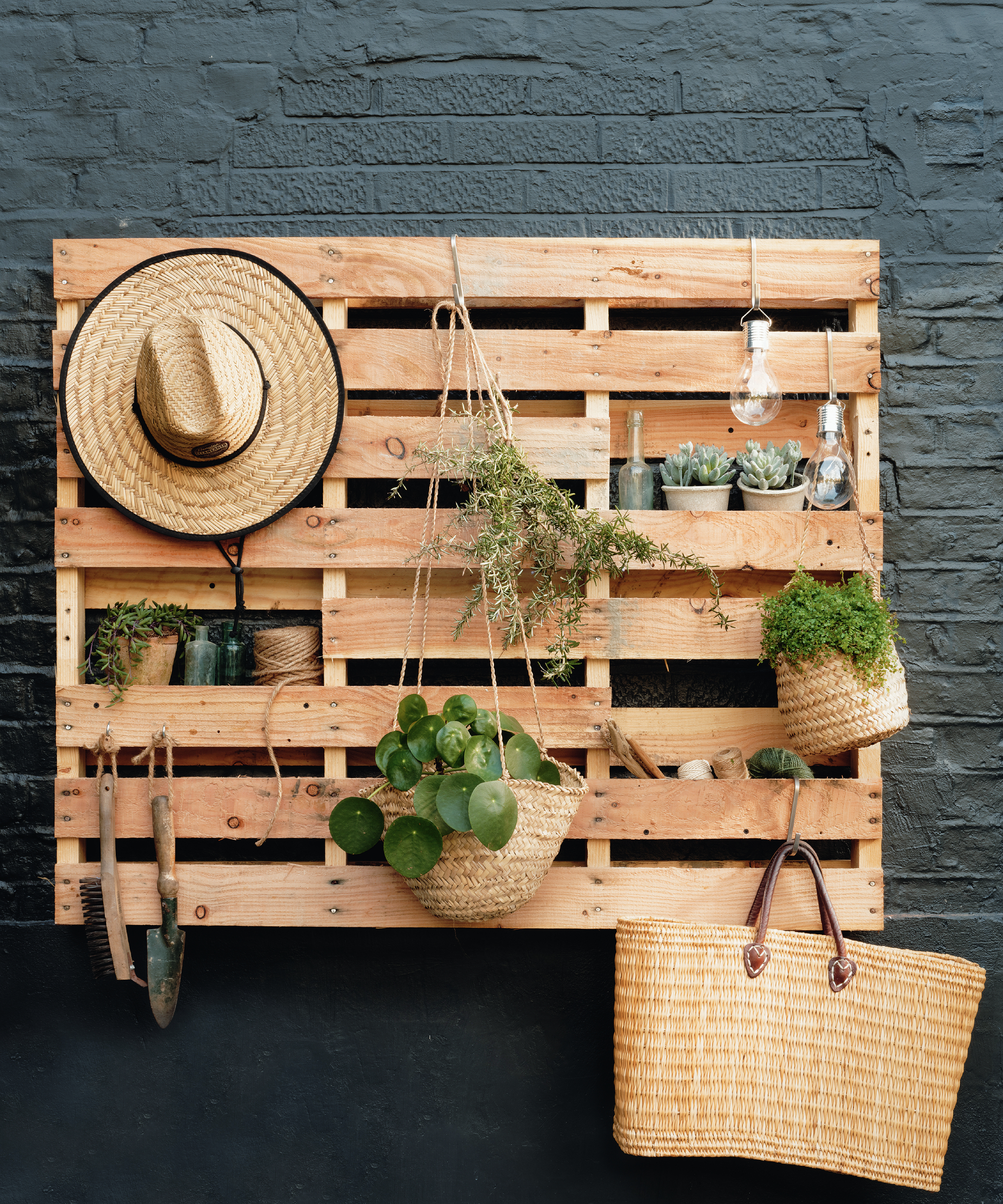 A set of wood shelves hanging on a wall, with plants in baskets, garden tools and a straw hat.