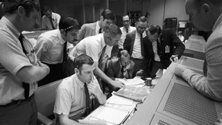 A group of flight controllers gathers around the console of Glenn S. Lunney
