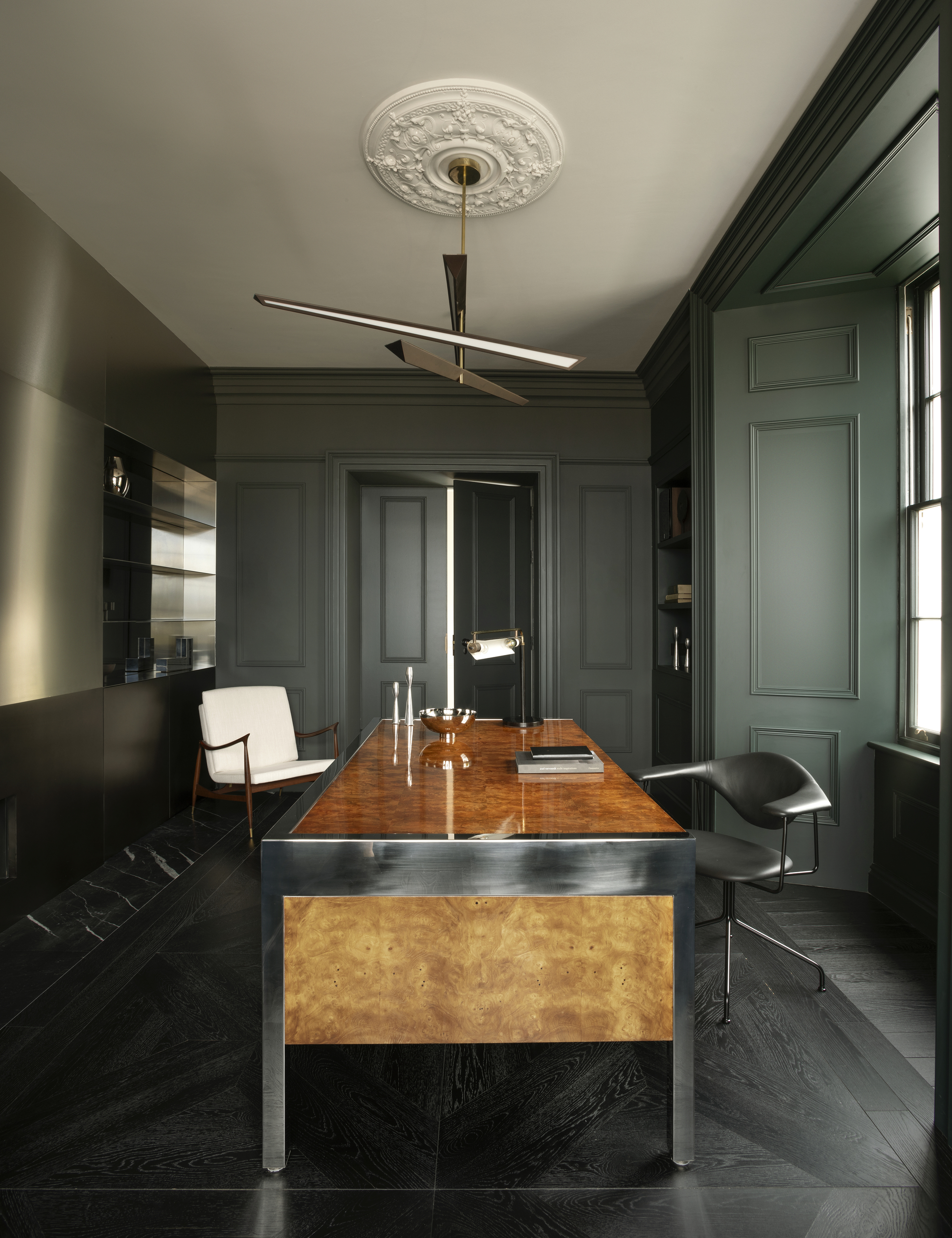 home office with green walls and paneling, white ceiling with ceiling rose and pendant light, burl and chrome desk, with black chairs and black timber floor