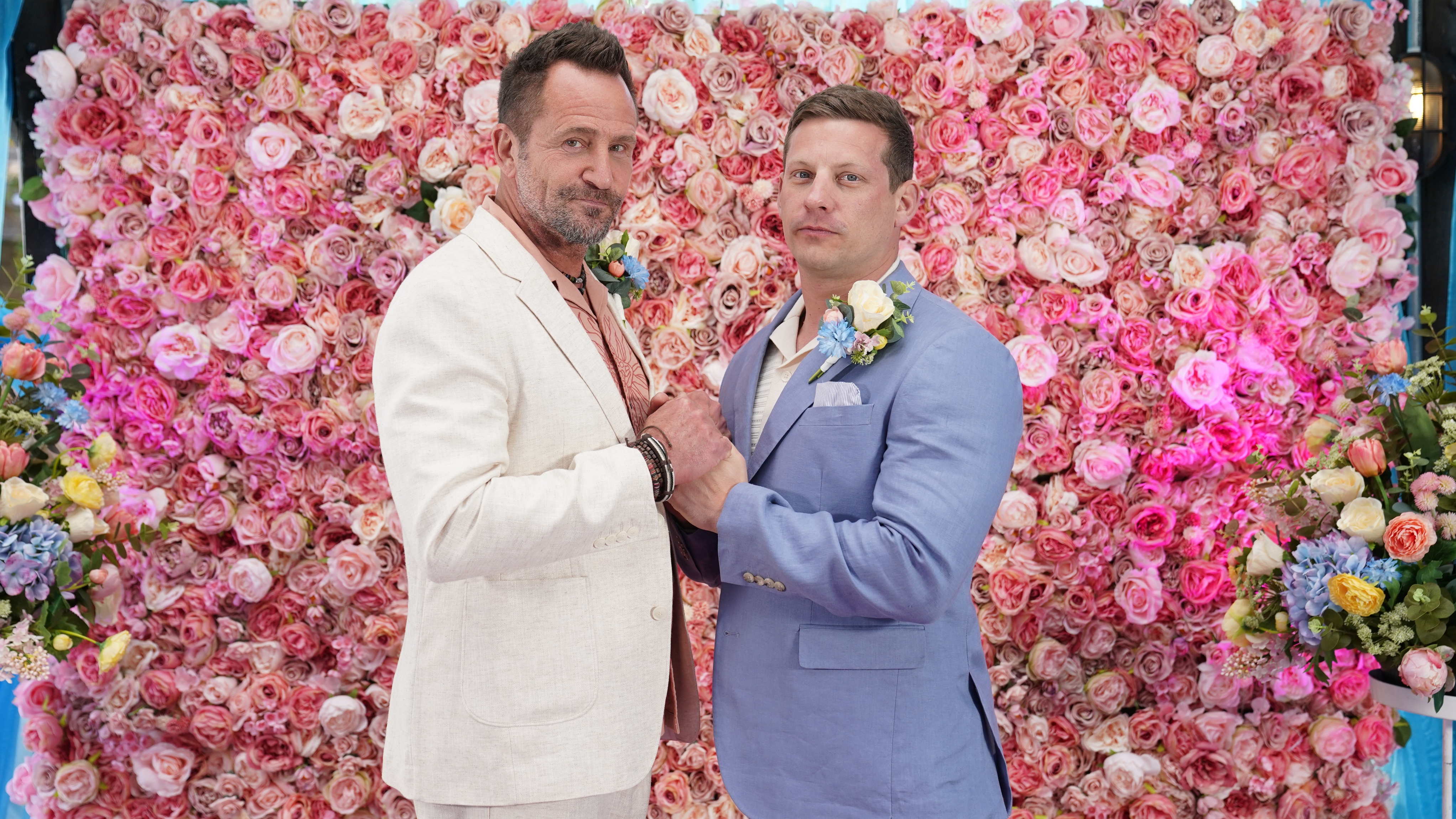 Jez and John Paul standing together in their wedding suits in front of a wall of roses.