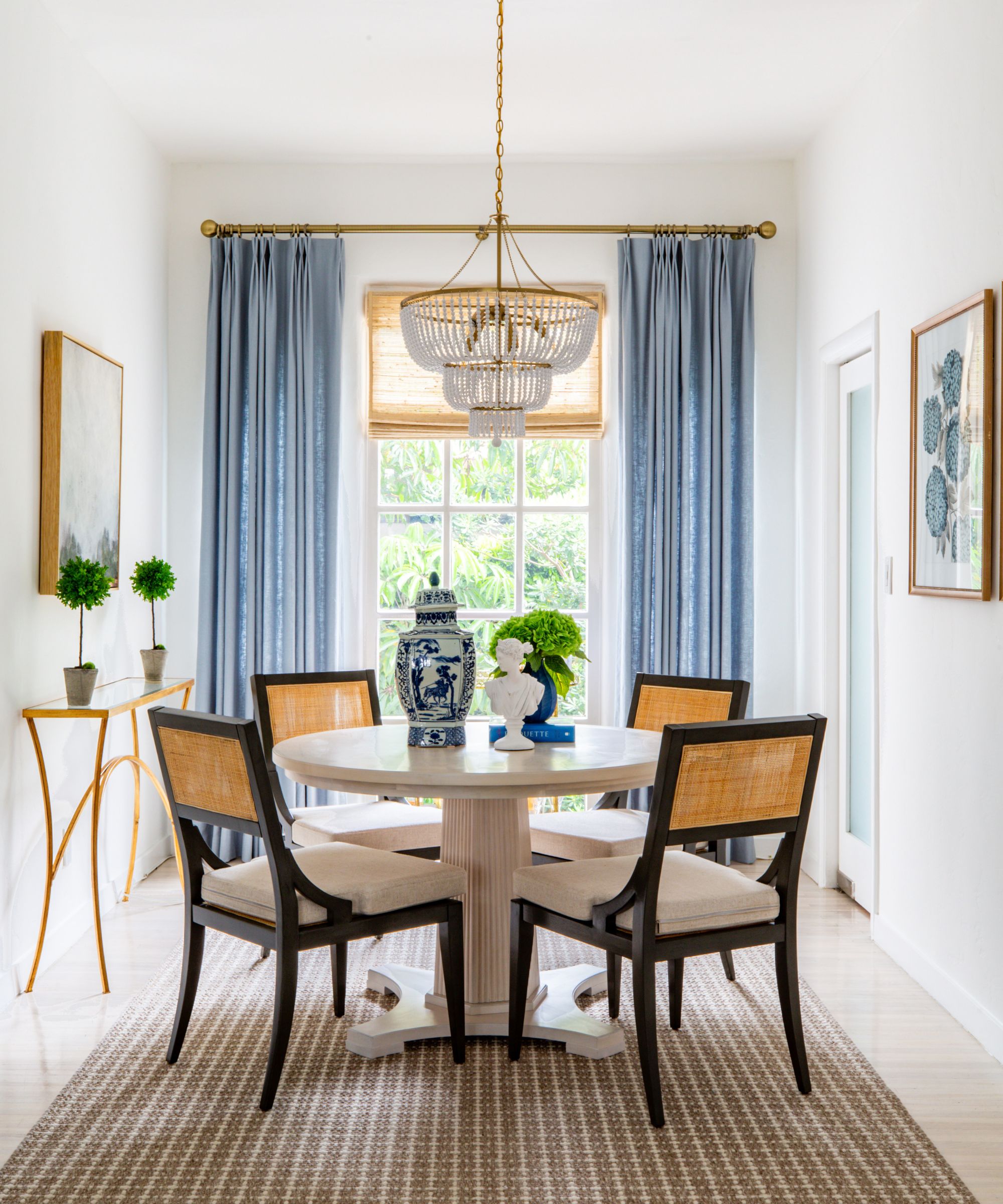 Dining room with white walls, brown patterned rug, a white round breakfast table with rattan dining chairs tucked underneath, blue long curtains and a chandelier.