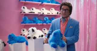Ty (Zach Galifianakis) at a toy expo in a pink booth, surrounded by stuffed plush Himalayan cats of various colours. He has a blue one on a podium in front of him, and he is combing its fur. He is looking off to the side and smirking.