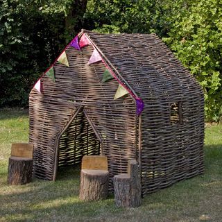 Children's Hazel Den, dark brown woven hazel with opening for door and window, colourful bunting at the top of the den and three wooden log chairs in front