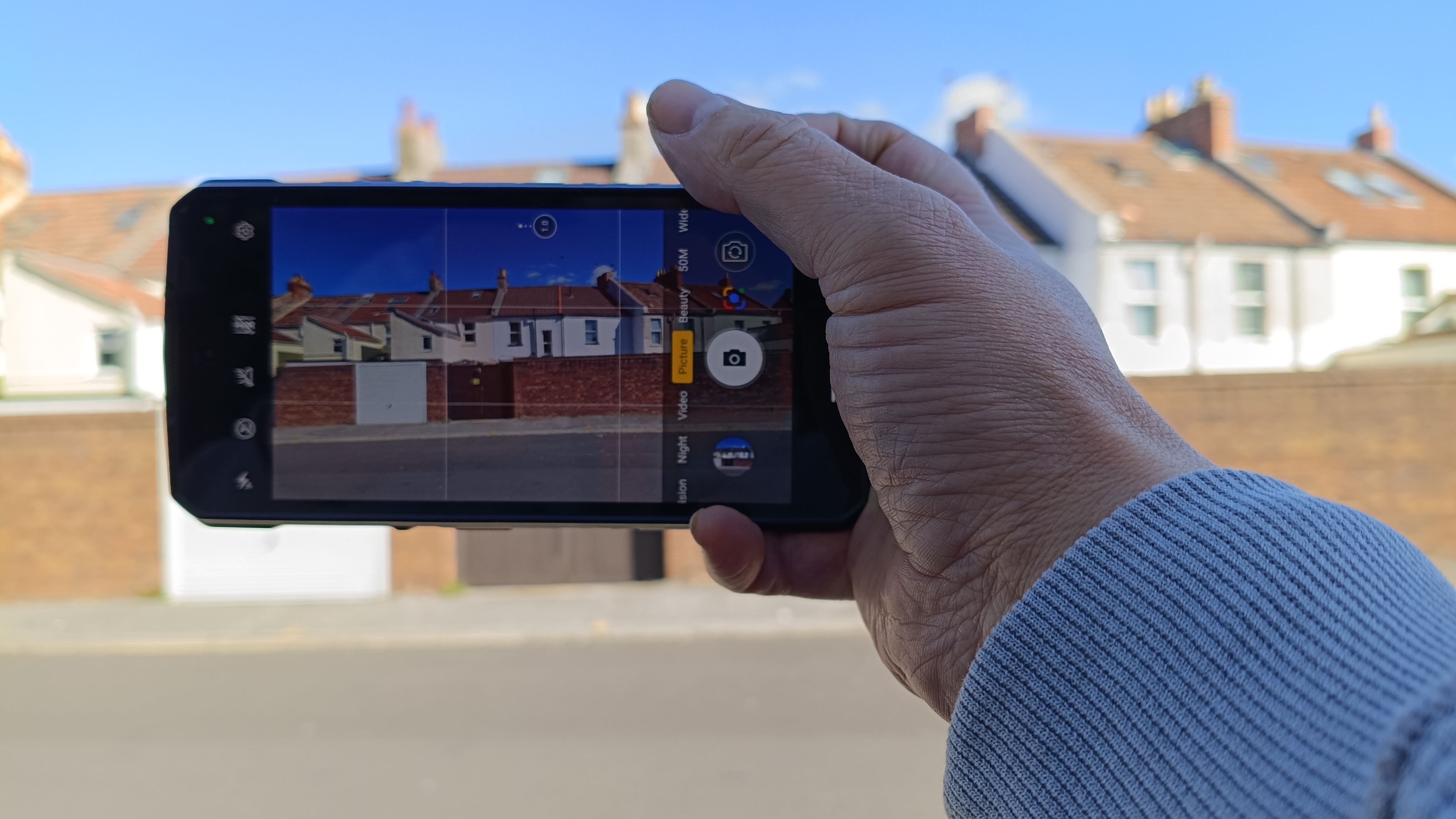 Hand holding Ulefone Armor 29 Ultra phone, with its camera app open, in front of a row of terraced houses under a sunny blue sky