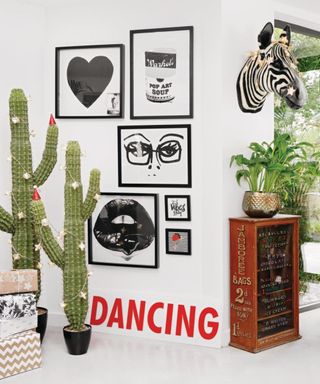 Detail of artwork on white walls, two llarge fake cactus with fairy lights, a zebra head on the wall and white ceramic tile floor. Sign saying Dancing. Edwardian family house in North London, home to Lucy St George and her partner Paul and their family.