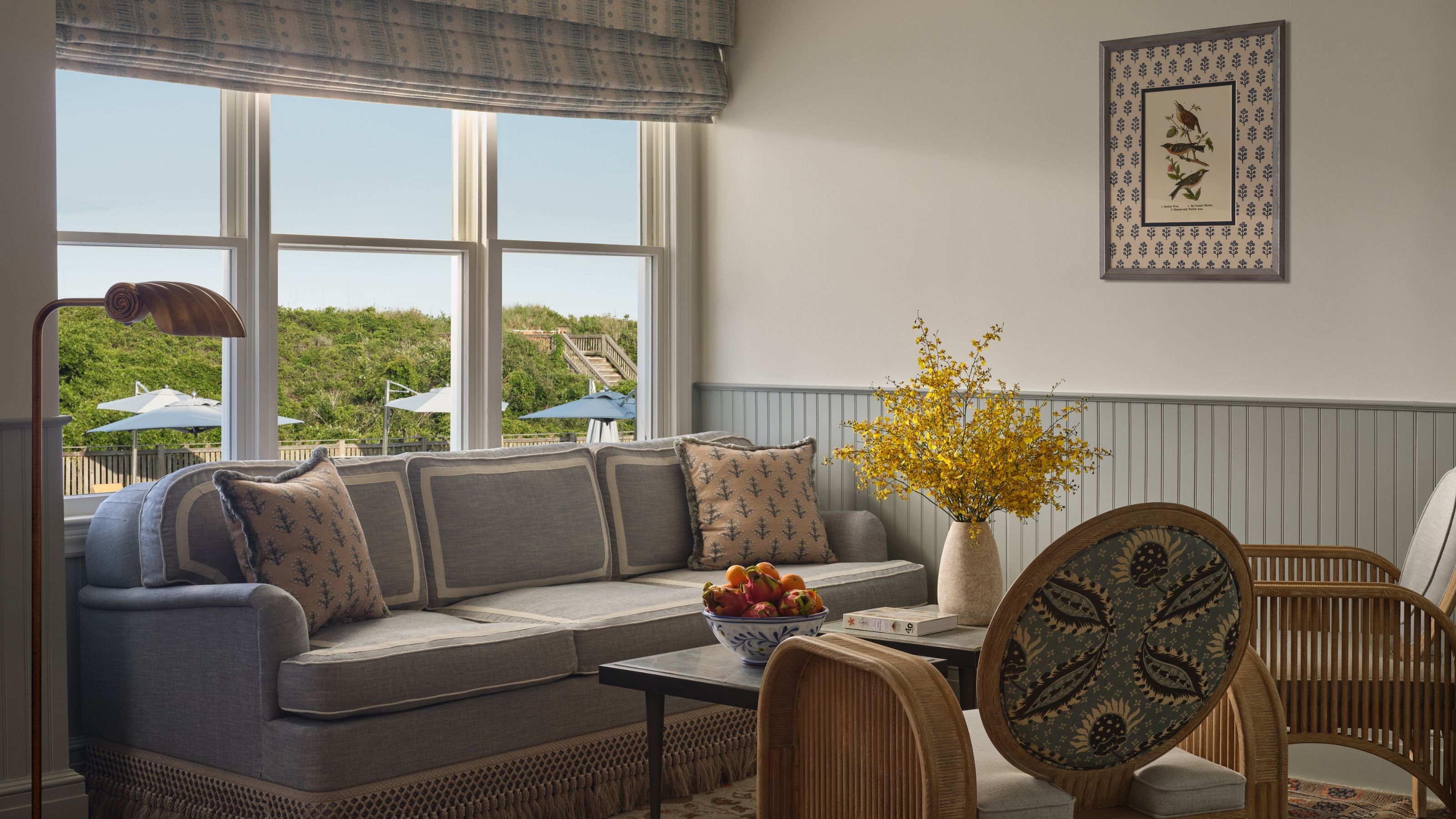 A bright sitting area featuring a grey sofa with patterned throw pillows next to a large window, complemented by a vase of yellow flowers on the coffee table.