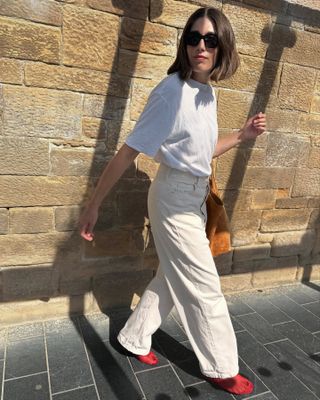 British influencer Lucy Alston wearing oversize sunglasses, a relaxed white T-shirt, camel Liffner suede tote bag, beige baggy jeans, and bright red mesh flats.