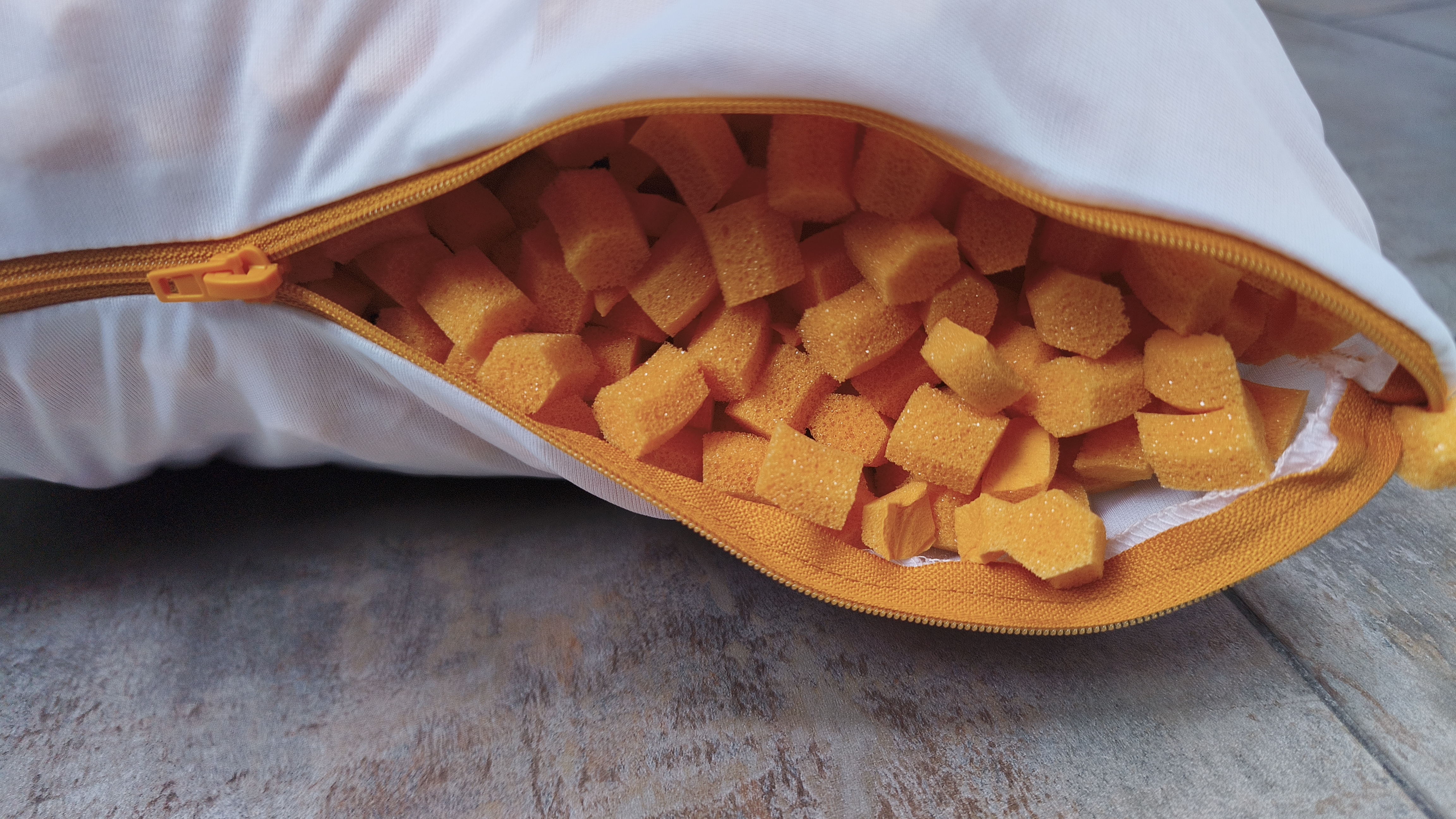 close-up view of an open orange zipper on a white pillowcase, revealing a dense filling of small, bright orange hexagonal foam pieces.