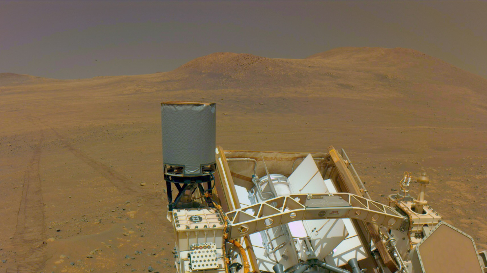 selfie taken by a mars rover, showing bits of its hardware in the foreground and rover tracks extending across a barren reddish-sand landscape in the background