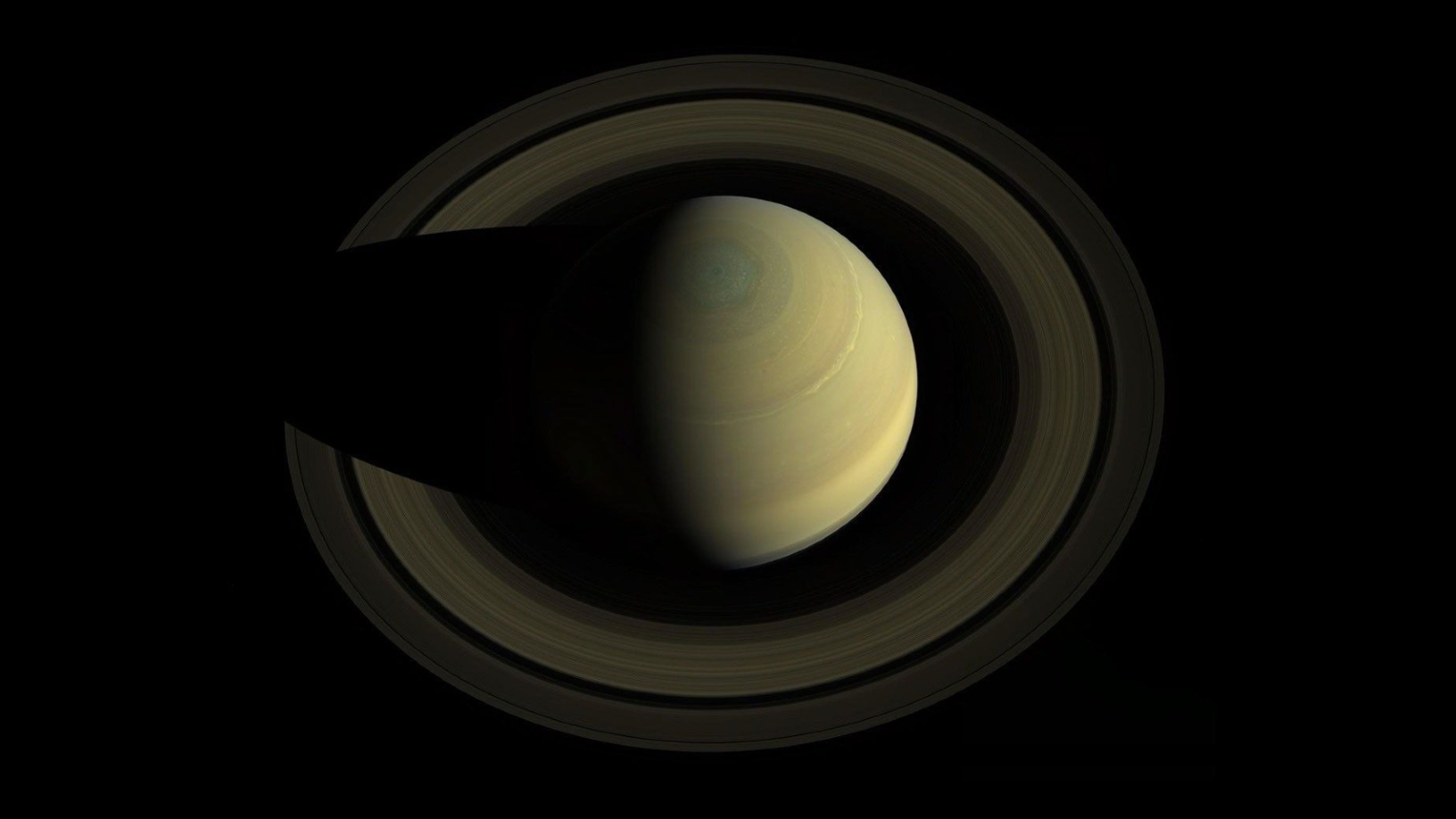 The planet Saturn is shown from above, shining against the blackness of space. Cloud bands line its cloud surface and a great hexagonal storm pattern is visible crowning its north polar region. A vast system of rings encircle the gas giant.
