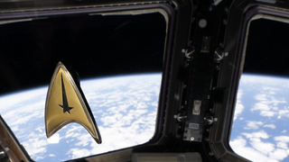 A golden Star Trek delta badge with a tall black star at its point floats in front of windows showing Earth from the ISS.