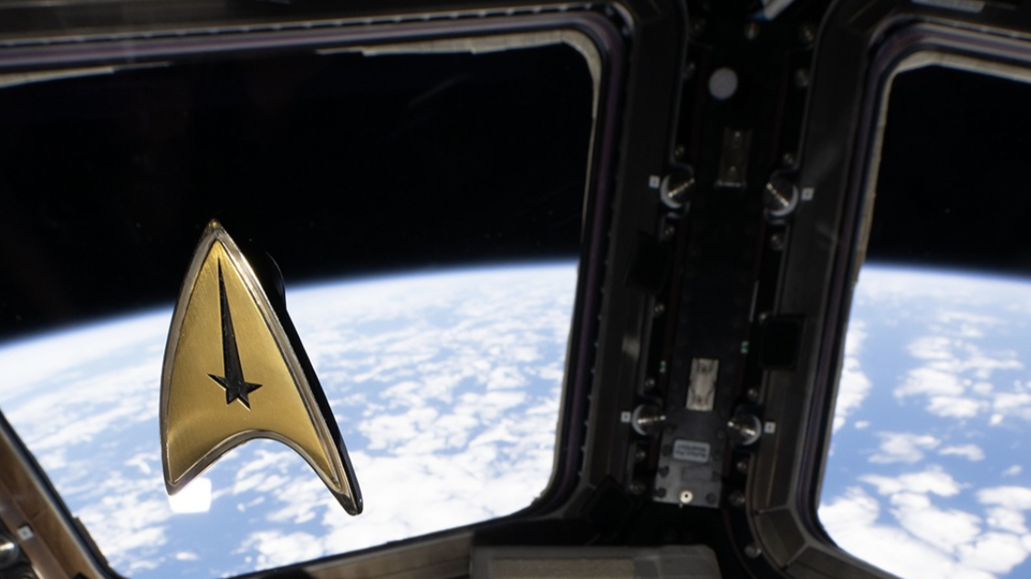 A golden Star Trek delta badge with a tall black star at its point floats in front of windows showing Earth from the ISS.