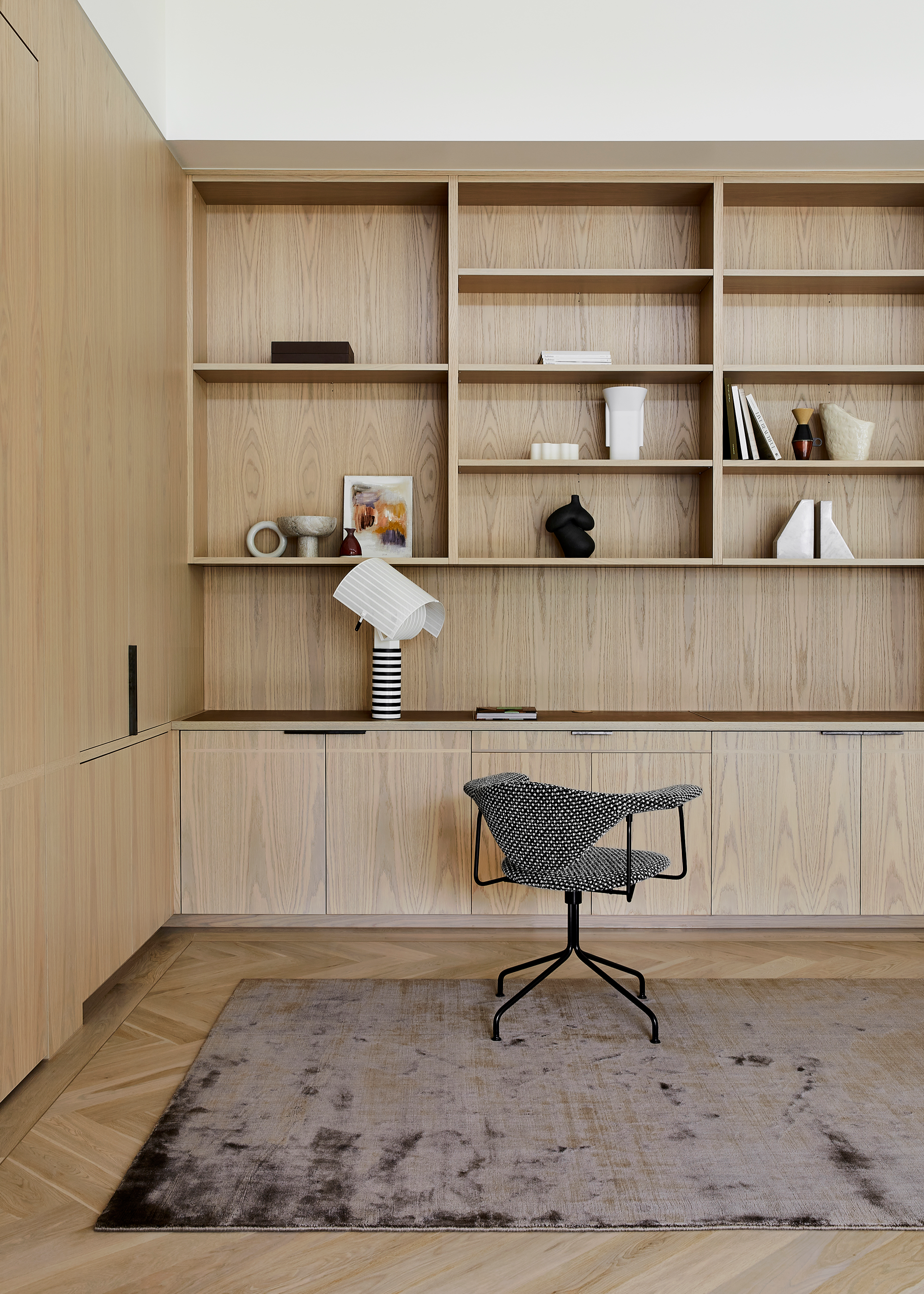A white study with a wood cupboard, in-built shelving, a swivel chair, and a deep purple rug