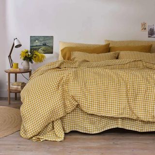 A large bed with a yellow gingham duvet and pillowcases and a circular wood bedside table holding a lamp