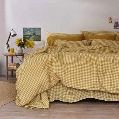 A large bed with a yellow gingham duvet and pillowcases and a circular wood bedside table holding a lamp