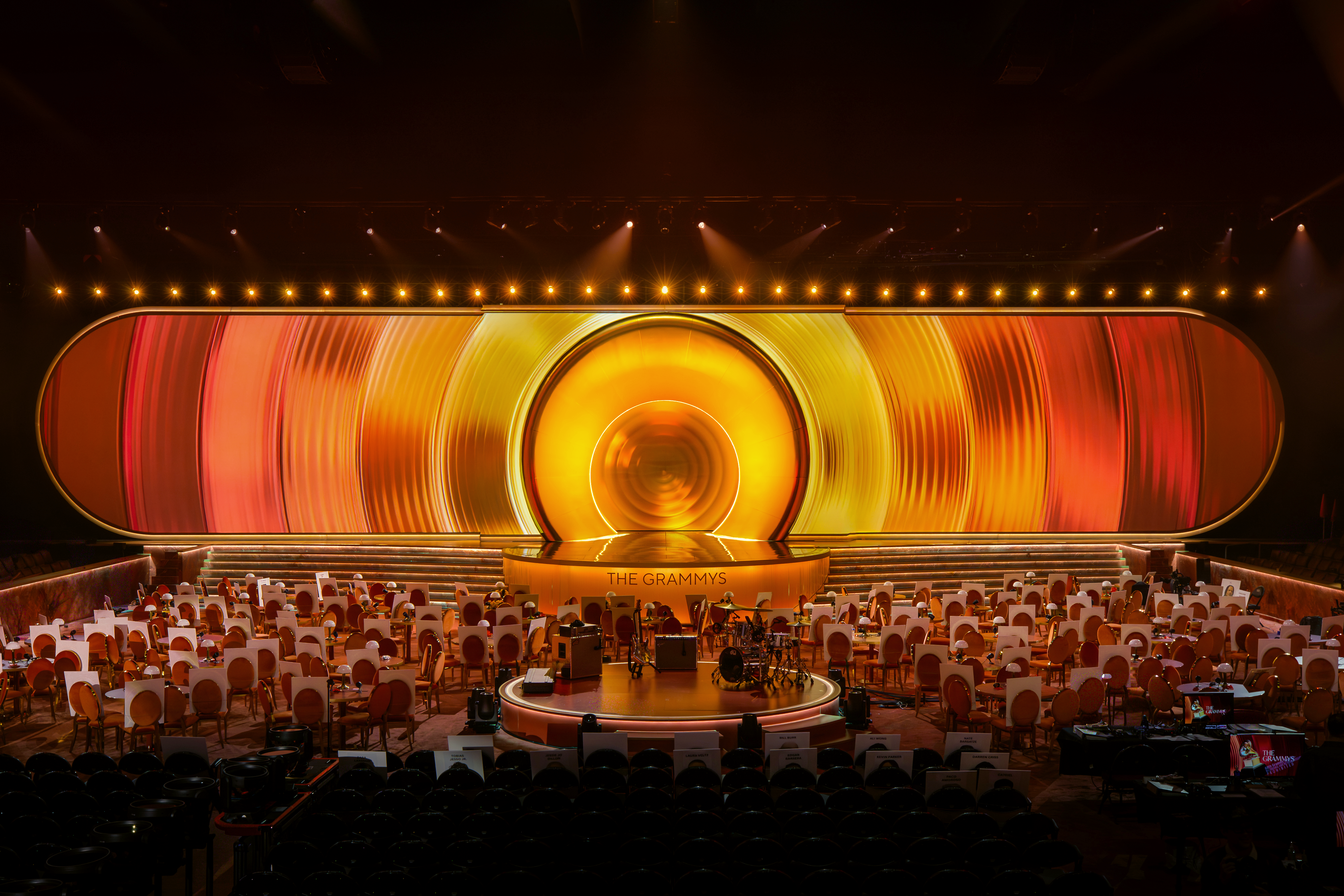An aerial view of a performance stage with a sculptural, abstract structure of orange, red, and yellow swathes, foregrounded by a series of ceremony round tables with lamps as centerpieces.