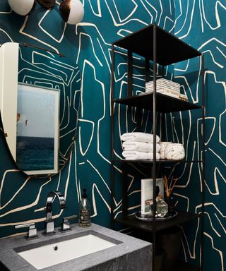Bathroom with bold, green botanical wallpaper, black metal shelving unit with towels and decorative items, oval style mirror above industrial, dark grey sink, wall light with two rounded opal glass diffusers
