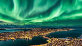 A cityscape illuminated by warm yellow lights sits below a stunning display of green northern lights sweeping across the night sky. Snow-covered mountains and a calm body of water add to the serene, wintery atmosphere.