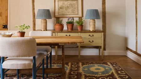 Dining room with a wooden painted console with two large gingham blue lamps on top, a wooden long dining table, upholstered dining chairs with a blue frame, and a brown and blue kilim rug beneath