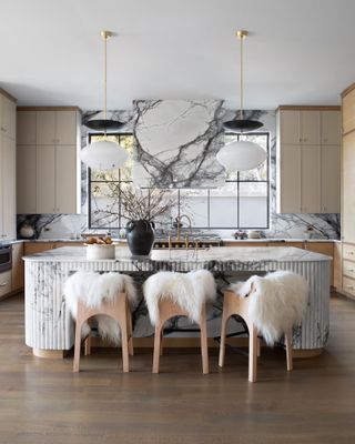 Kitchen with beige cabinetry, veined marble splashback and cooker hood, and round fluted marble island with wood bar stools