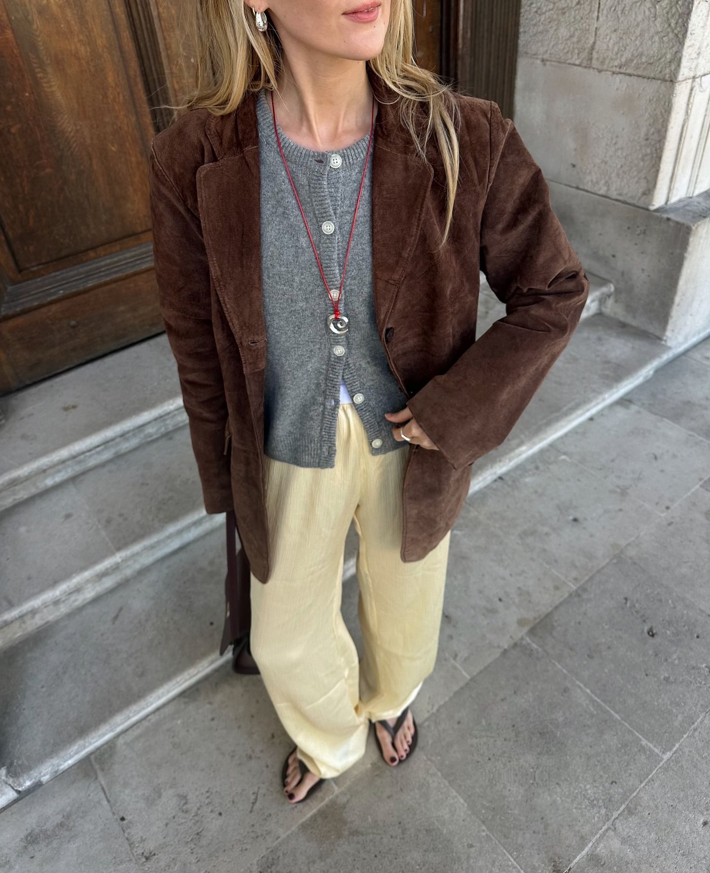 British style influencer Chloe Butler poses on a London sidewalk wearing silver chunky earrings, a brown suede jacket, gray cardigan sweater, a red cord necklace with a silver spiral pendant, yellow satin pants, and black flip-flop sandals