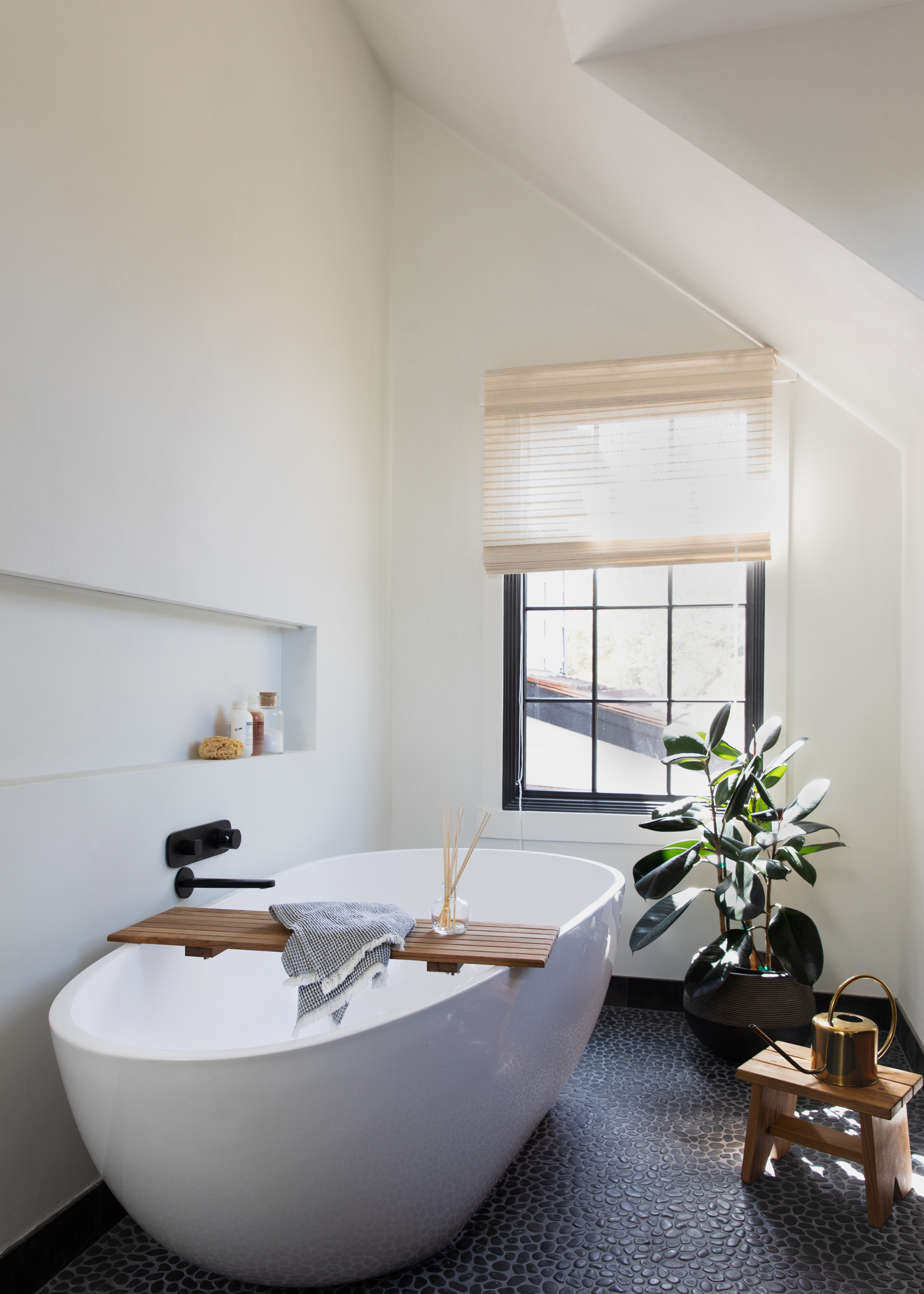 A tranquil bathroom with a standing tub, a sunlit window, a potted plant and a wooden stool with a watering can.