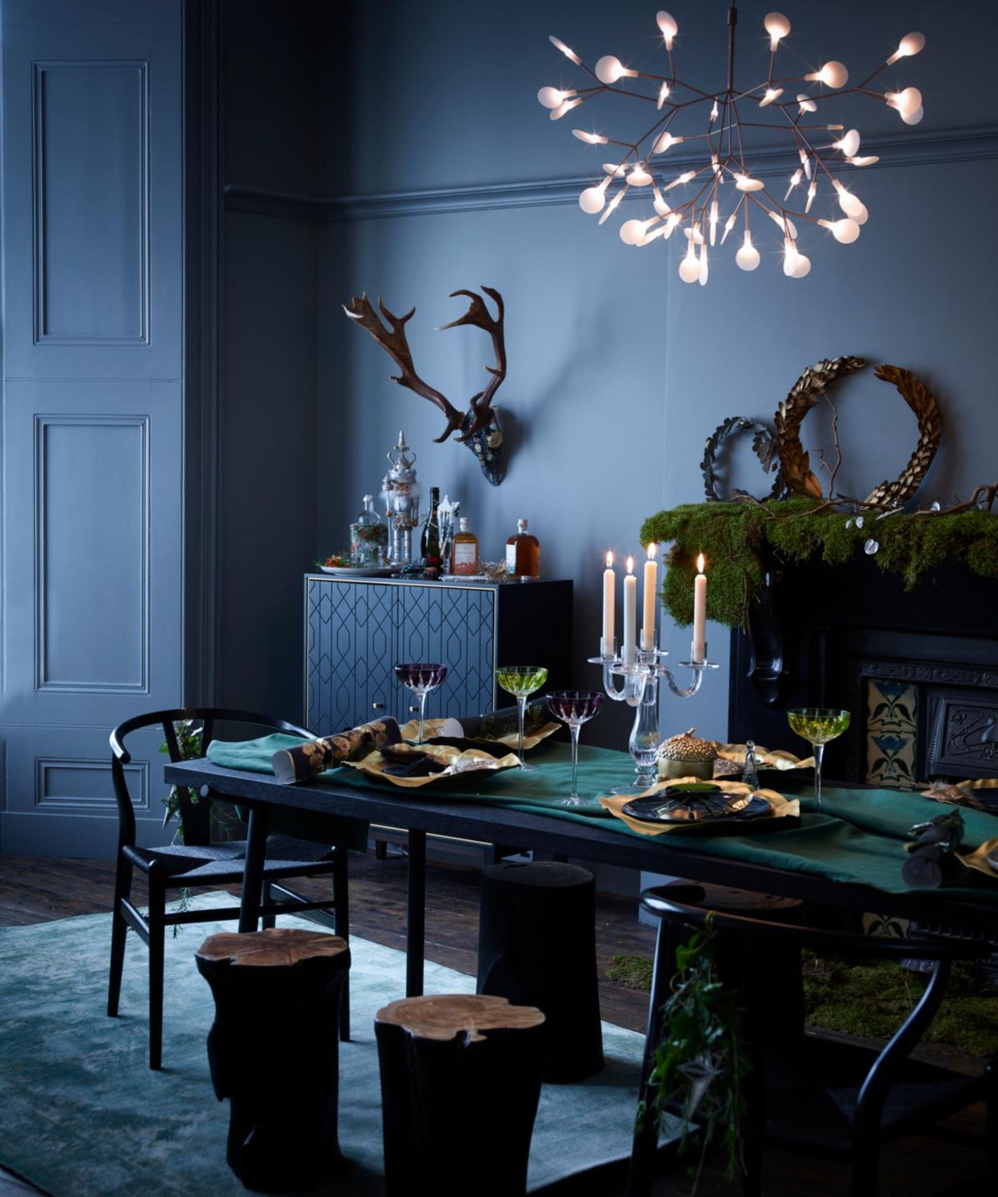 A dining room table set for a Christmas meal, with table settings, blue plates and gold table decorations, a chandelier, stags head on the wall and a mantlepiece with foliage decorations.