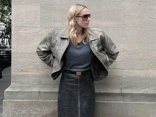 Elly wears a black top and black leather skirt with a brown. jacket while turning head and wearing sun glasses