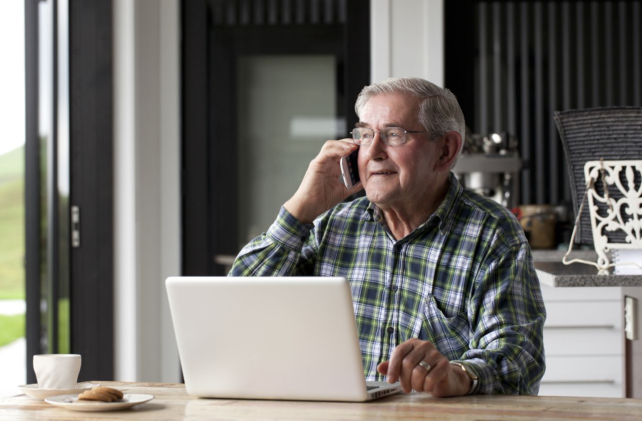 Senior male talking on smartphone while seated at table. Laptop is on table in front of him.