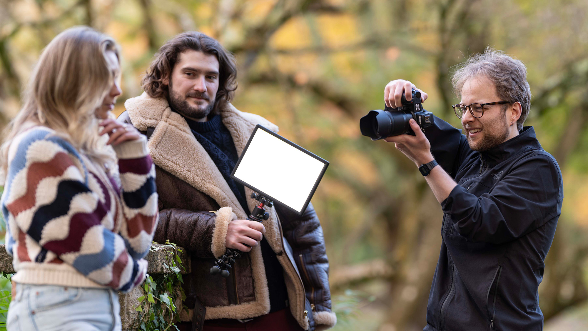 Photographer photographs model with second model off to one side holding LED light panel, against a backdrop of trees