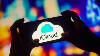 iCloud logo appearing on a smartphone being held in outstretched arms by a silhouetted person against a multicoloured background