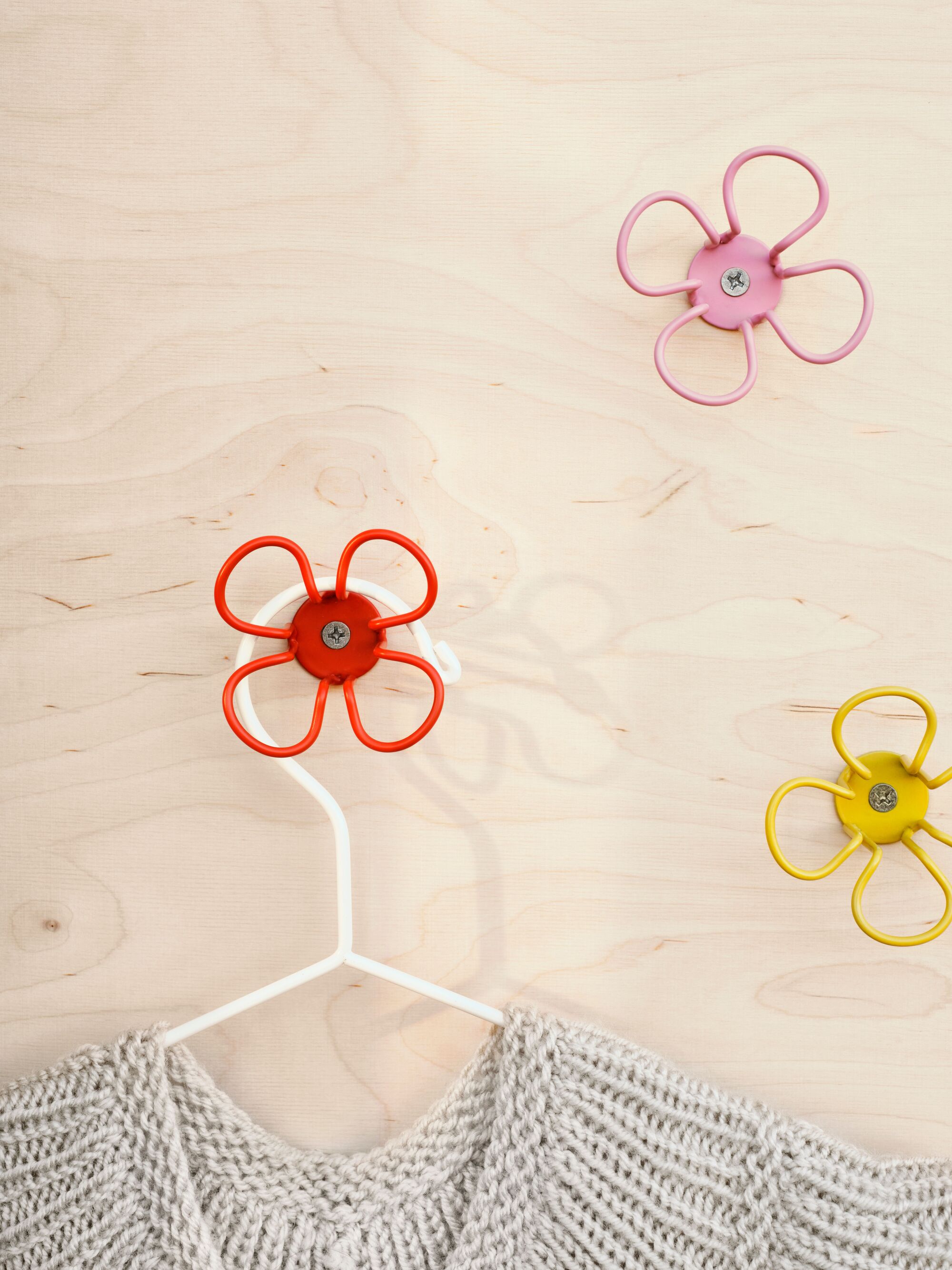Close-up image of a light wooden wall with three colorful, metal flower-shaped clothing hooks. There is a gray seater hanging off of one of them.