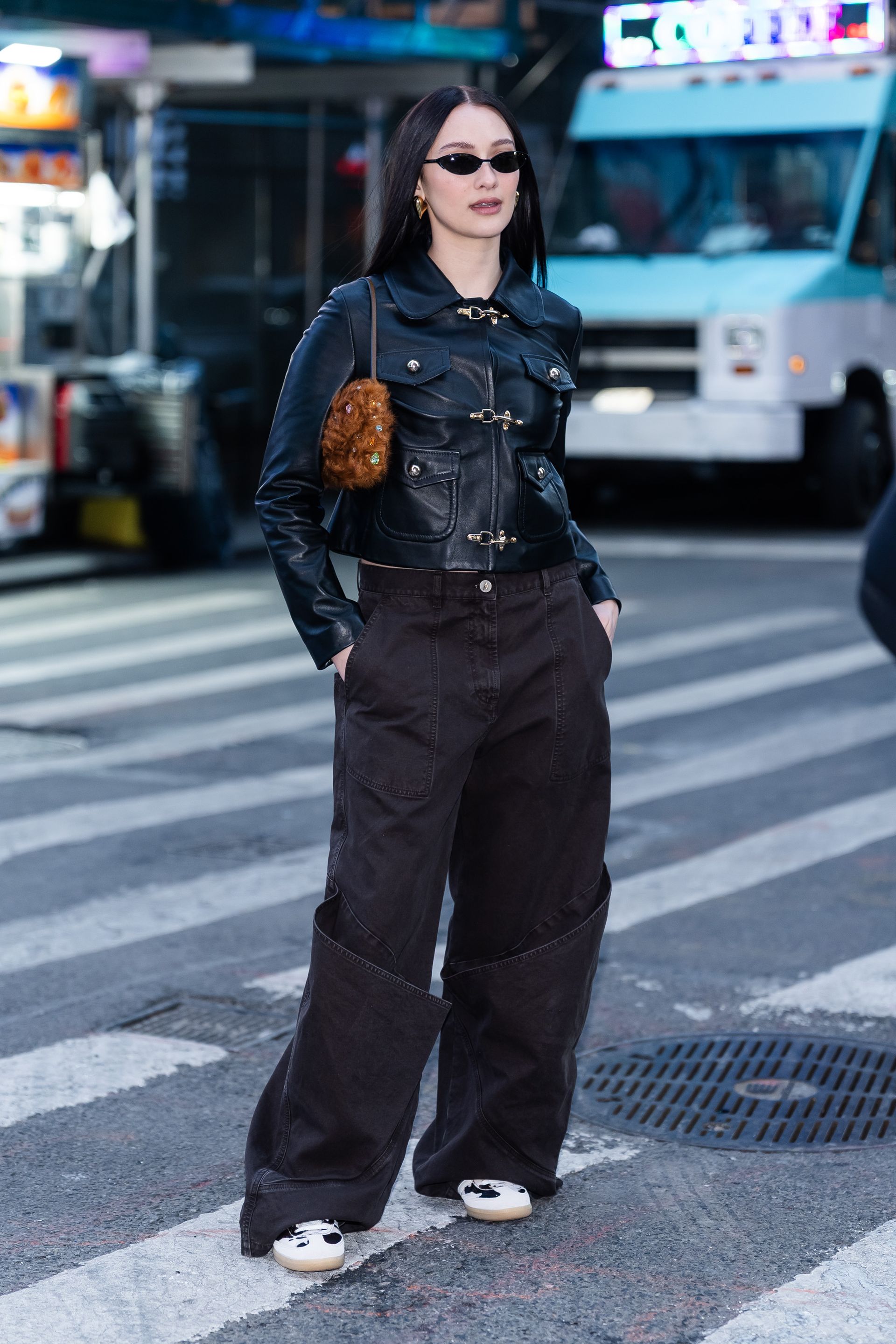 Lola Tung stands on the road in New York wearing a leather toggle jacket with low-rise jeans, cow print trainers and a furry bag.