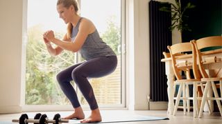 Woman lowering into a squat with dumbbells on her yoga mat in living room