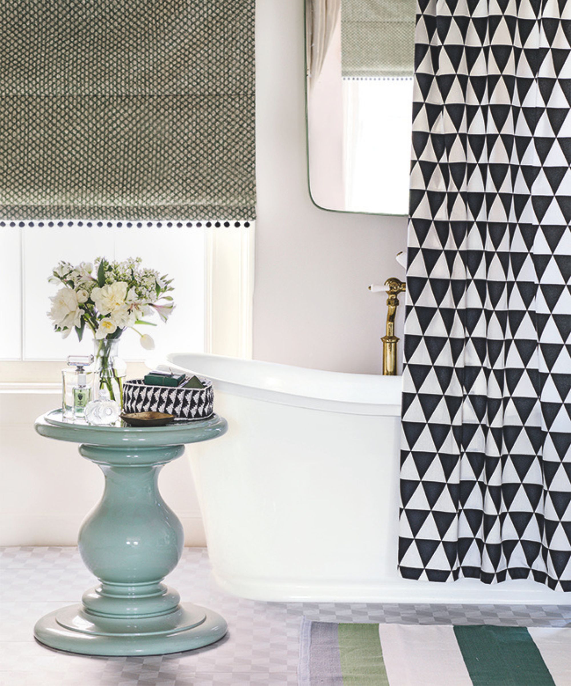 A white bathtub in a bright bathroom with a black and white shower curtain and gold faucet, mirror above, and a blue circular table with a clear vase of white flowers on top. Behind is a large window with green blind, and on the floor is a green and white striped rug.
