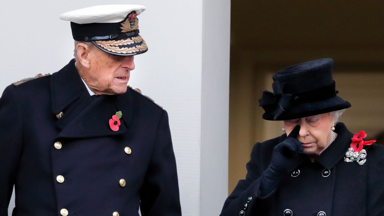 Queen Elizabeth wiping a tear while wearing a black coat and hat standing next to Prince Philip in a black coat and naval hat