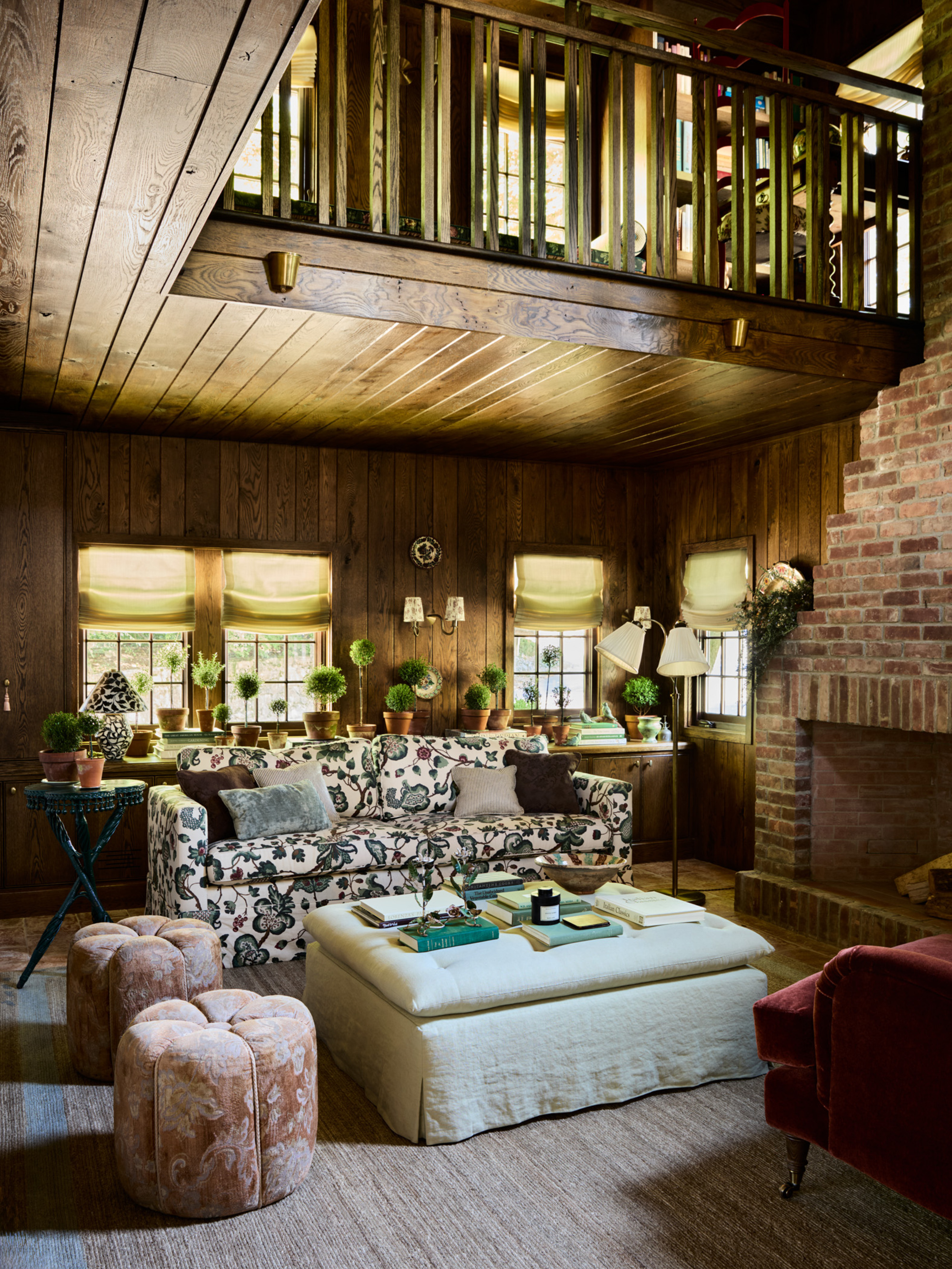 A wood-panelled living room with a loft above it. There is a patterned sofa, two pink pouffes, and a large, square, white linen-upholstered coffee table in the sitting area.