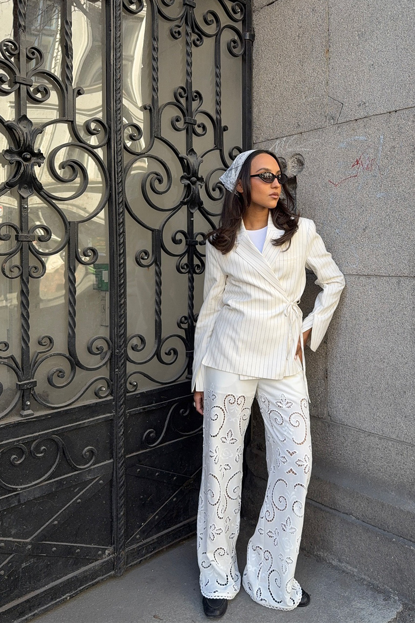 How to wear a bandana is shown in a photo of a woman standing in a doorway outside wearing a lace kerchief, black sunglasses, white striped blazer, white tank top, white eyelet trousers, and black loafers