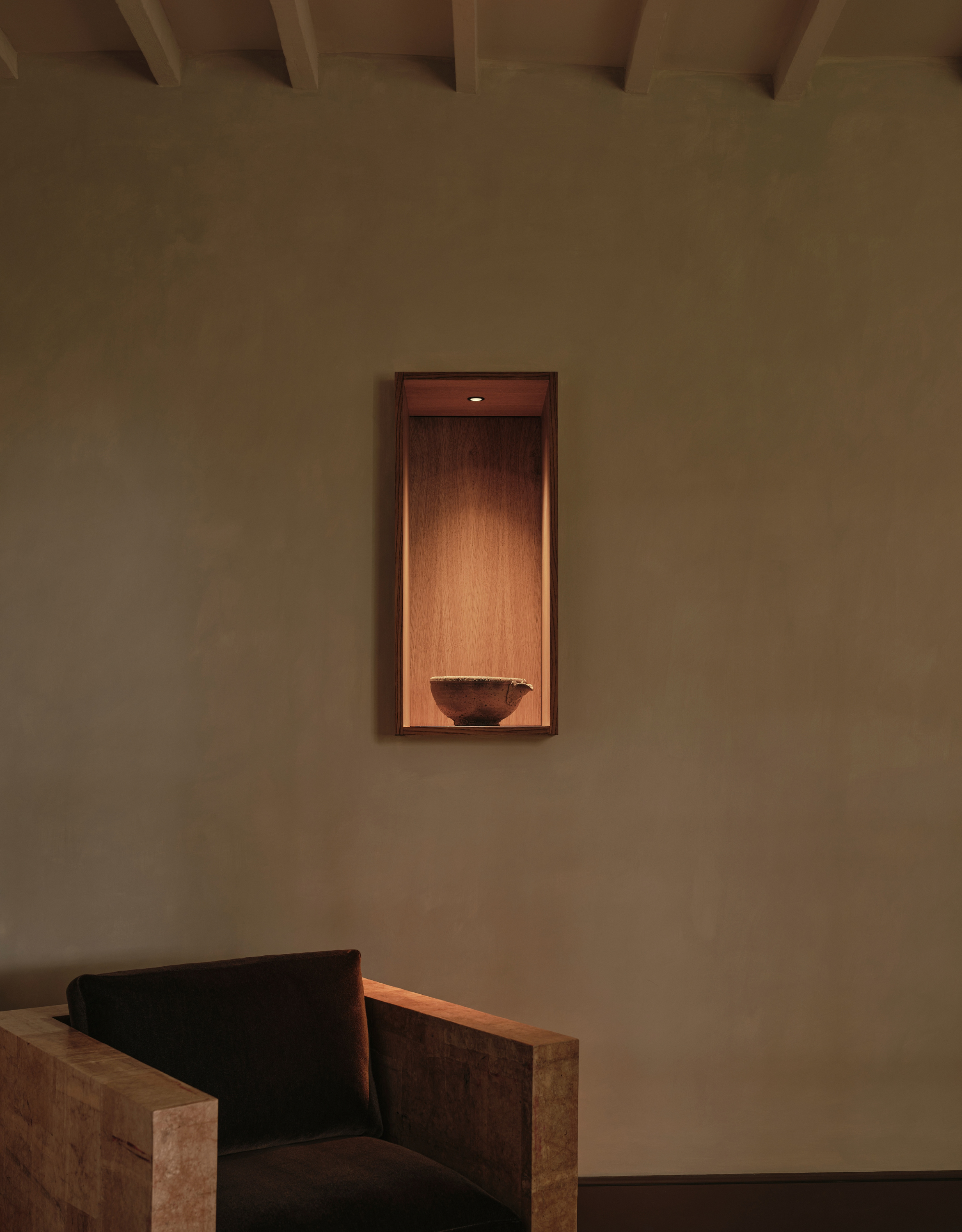 A display nook with a single spotlight over a ceramic bowl in a living room with a structured chair