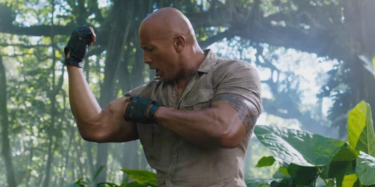 Dr. Xander Bravestone (The Rock) looks shocked at the size of his bicep as he flexes it in &#039;Jumanji: