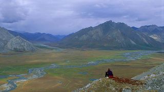A person seated and looking over a glacier valley in the Alaskan tundra with mountains in the background