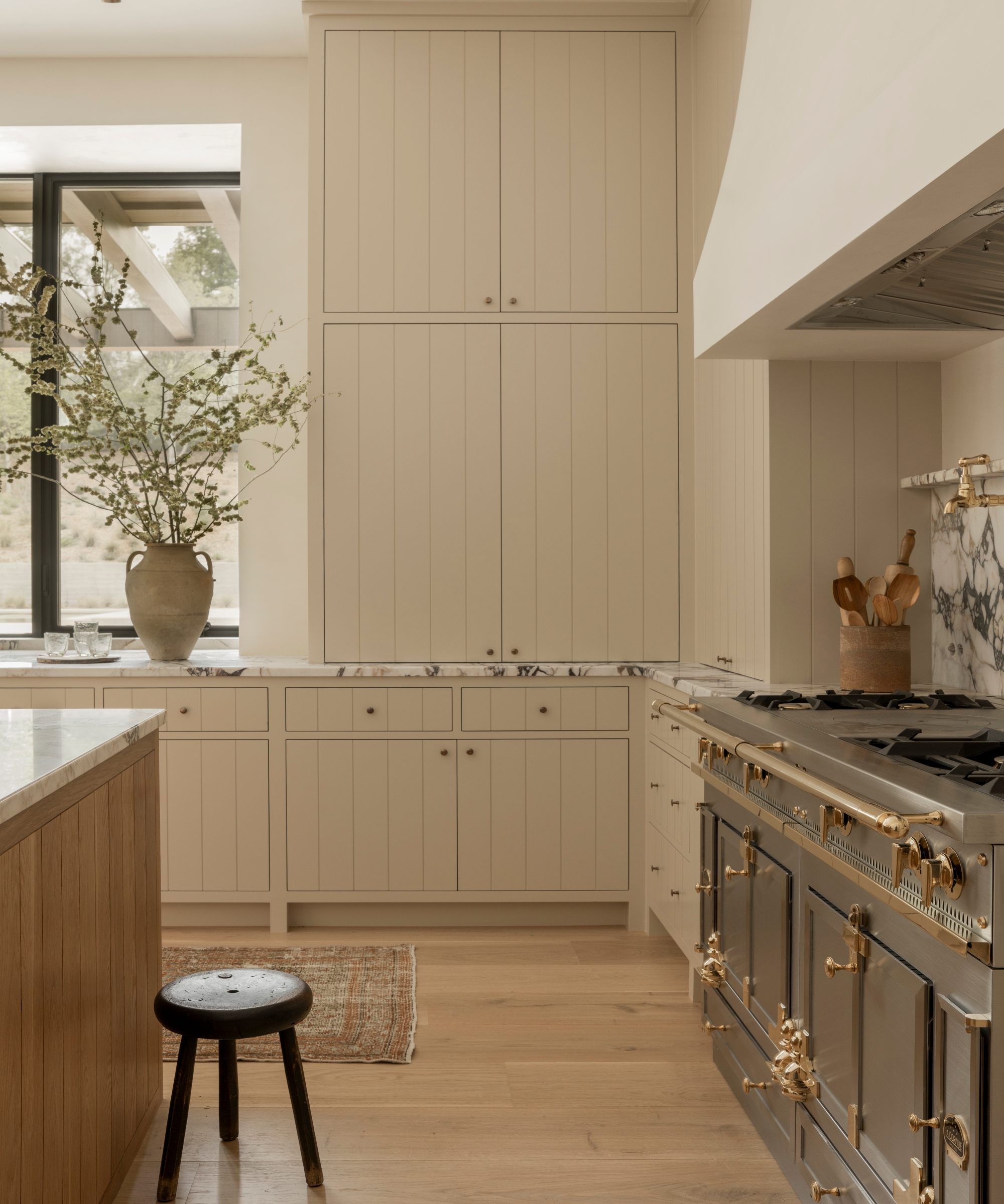 A warm neutral kitchen with taupe cabinets, a wood island, marble countertops, and black range cooker with brass accents